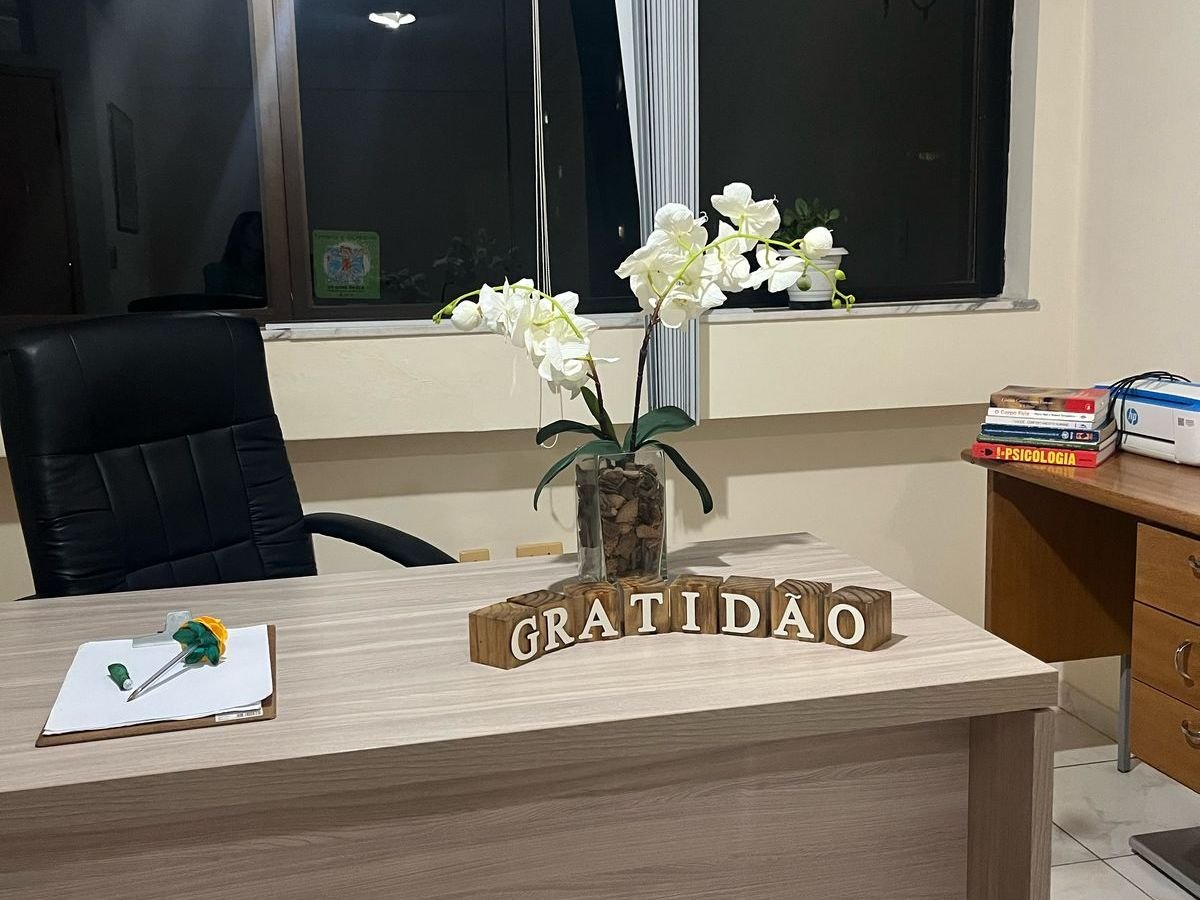 Mesa do consultório junto à janela, com orquídeas e uma estante de livros de psicologia ao fundo