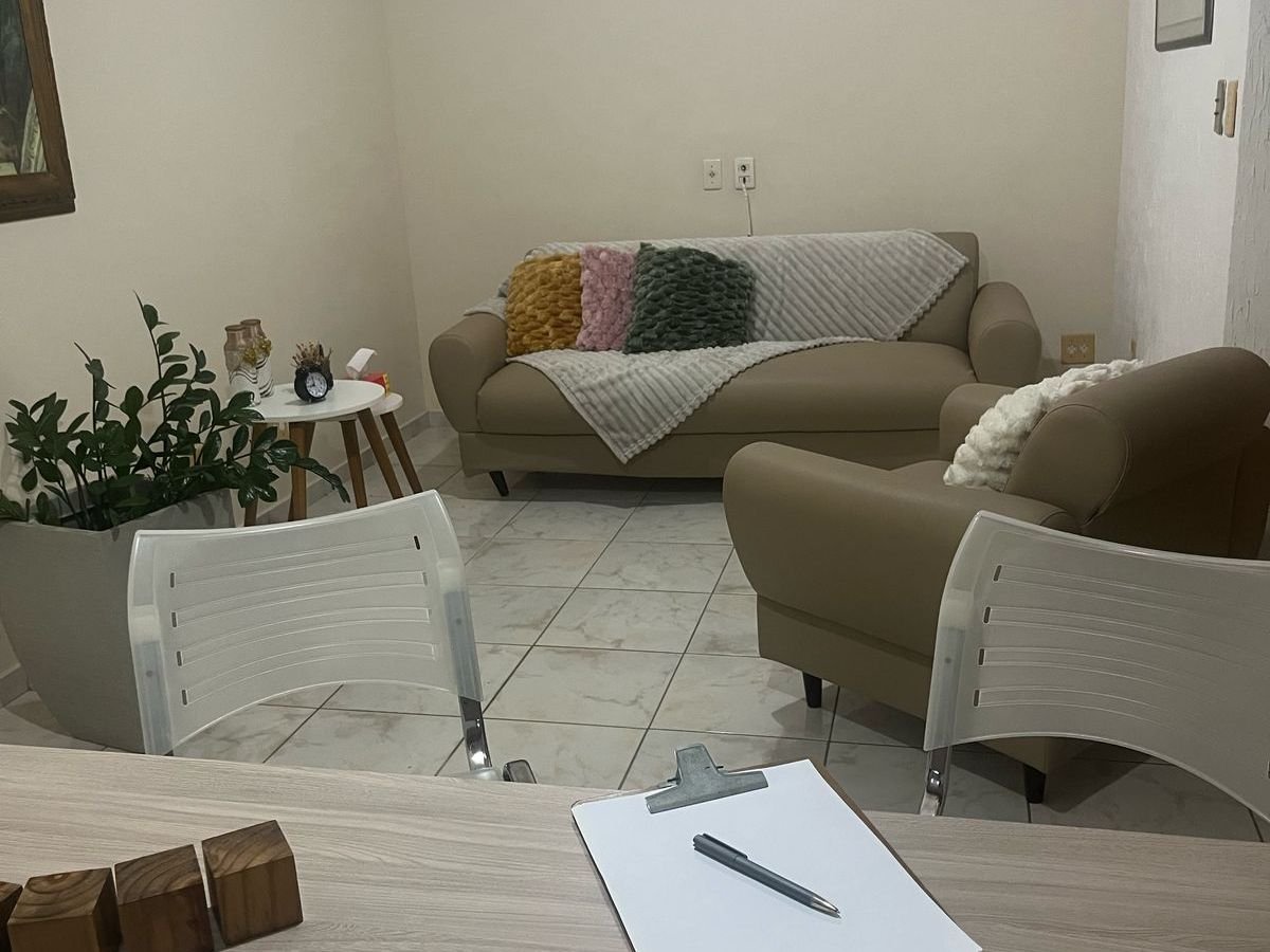 Sala de atendimento com sofá, poltrona, plantas e duas cadeiras diante de uma mesa